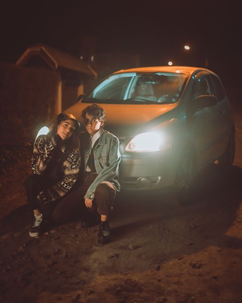 Free Full body of teenage girlfriends looking at camera while hunkering down near automobile with headlights in suburb area in evening time Stock Photo