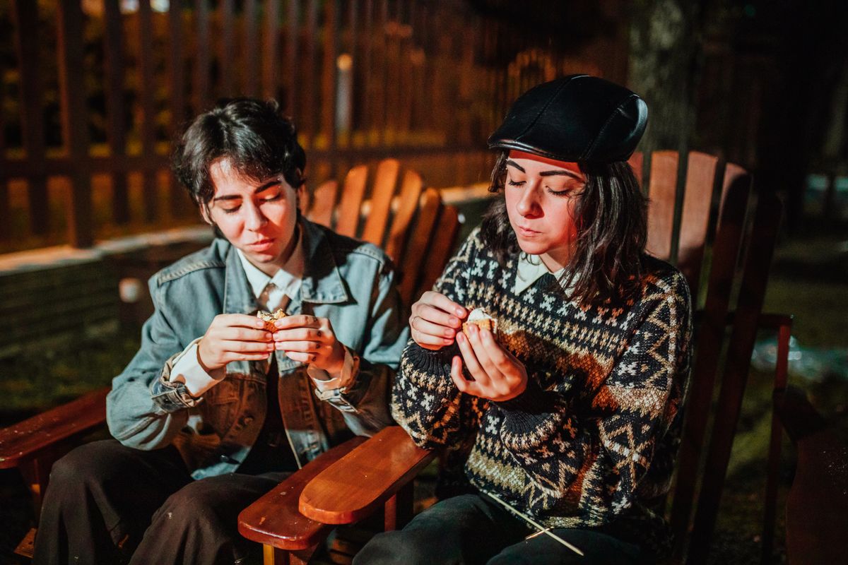 Teen females in casual wear eating sweet marshmallow in wooden chairs while spending time together in countryside in evening time