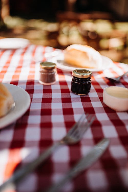 Free Small jars with sweet jam placed on table with checkered tablecloth and cutlery near plates in garden on blurred background Stock Photo