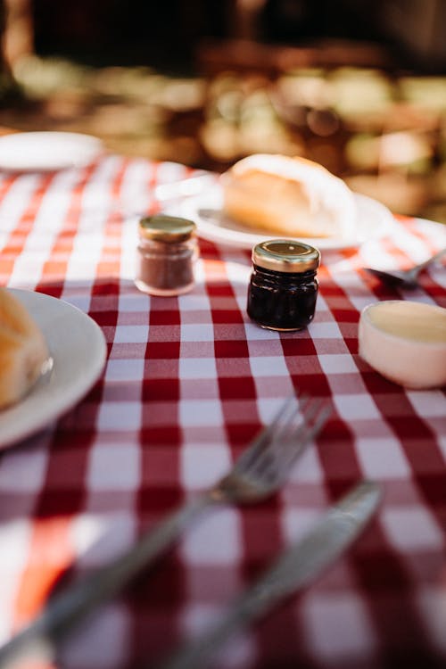 Small jars with sweet jam placed on table with checkered tablecloth and cutlery near plates in garden on blurred background