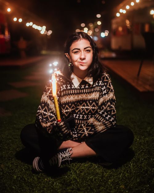 Free Full body of young female with fountain firework in hand looking at camera while sitting on grassy lawn in evening time in city Stock Photo