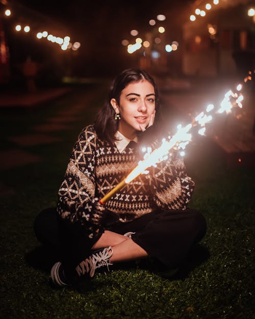Full body of young female in casual clothes looking at camera while sitting on lawn with sparkler candle on dark street