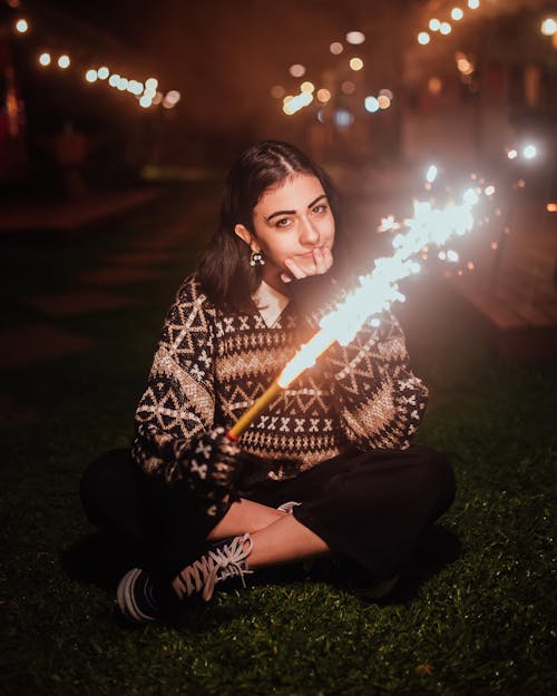 Full body of female in casual wear with fountain firework in hand looking at camera while sitting on street in evening time