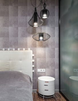 Comfortable bed with soft headboard placed at wall near white bedside table in stylish bathroom with hanging lamps in apartment