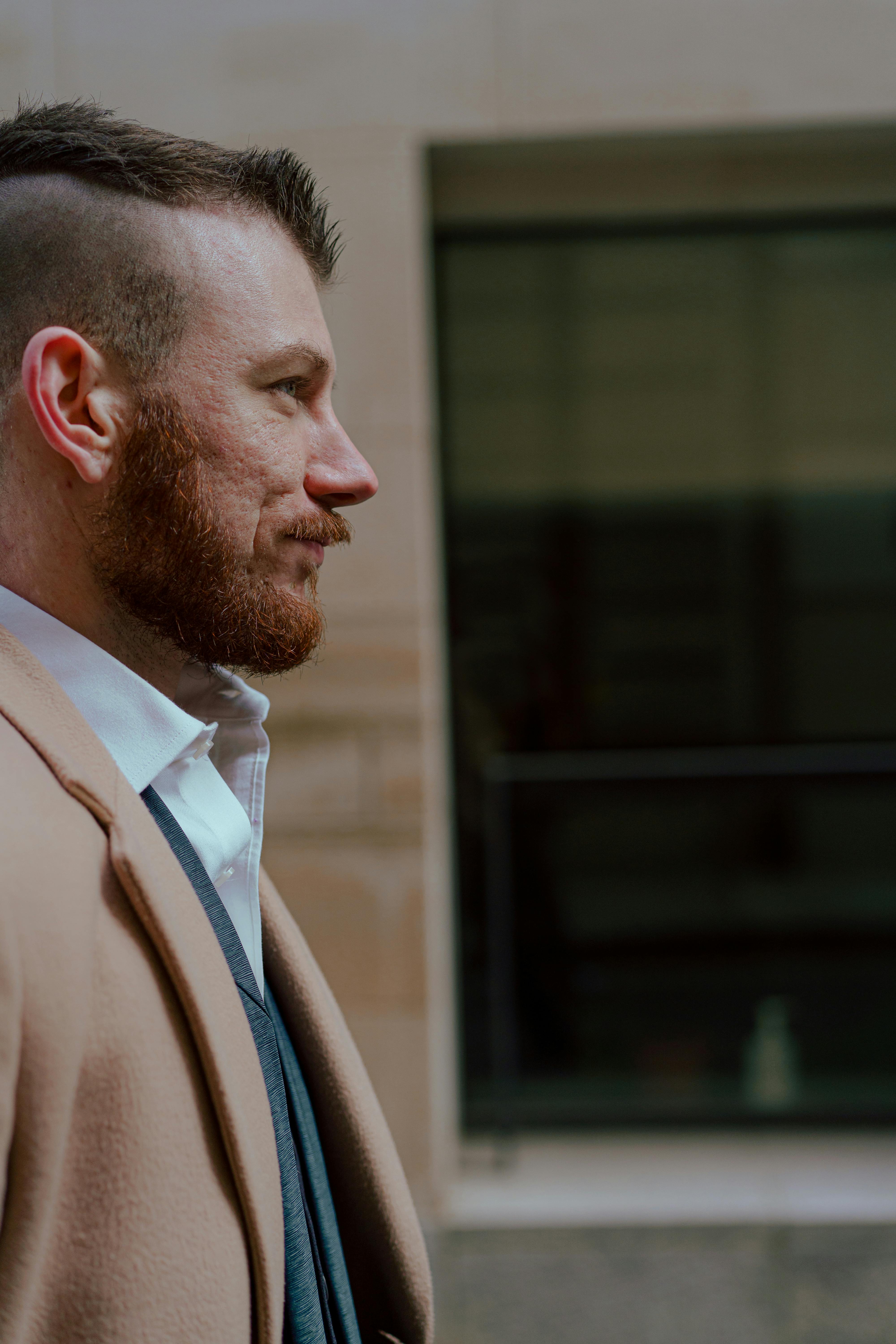 Side view portrait of a young man with beard and brown suit outdoors.