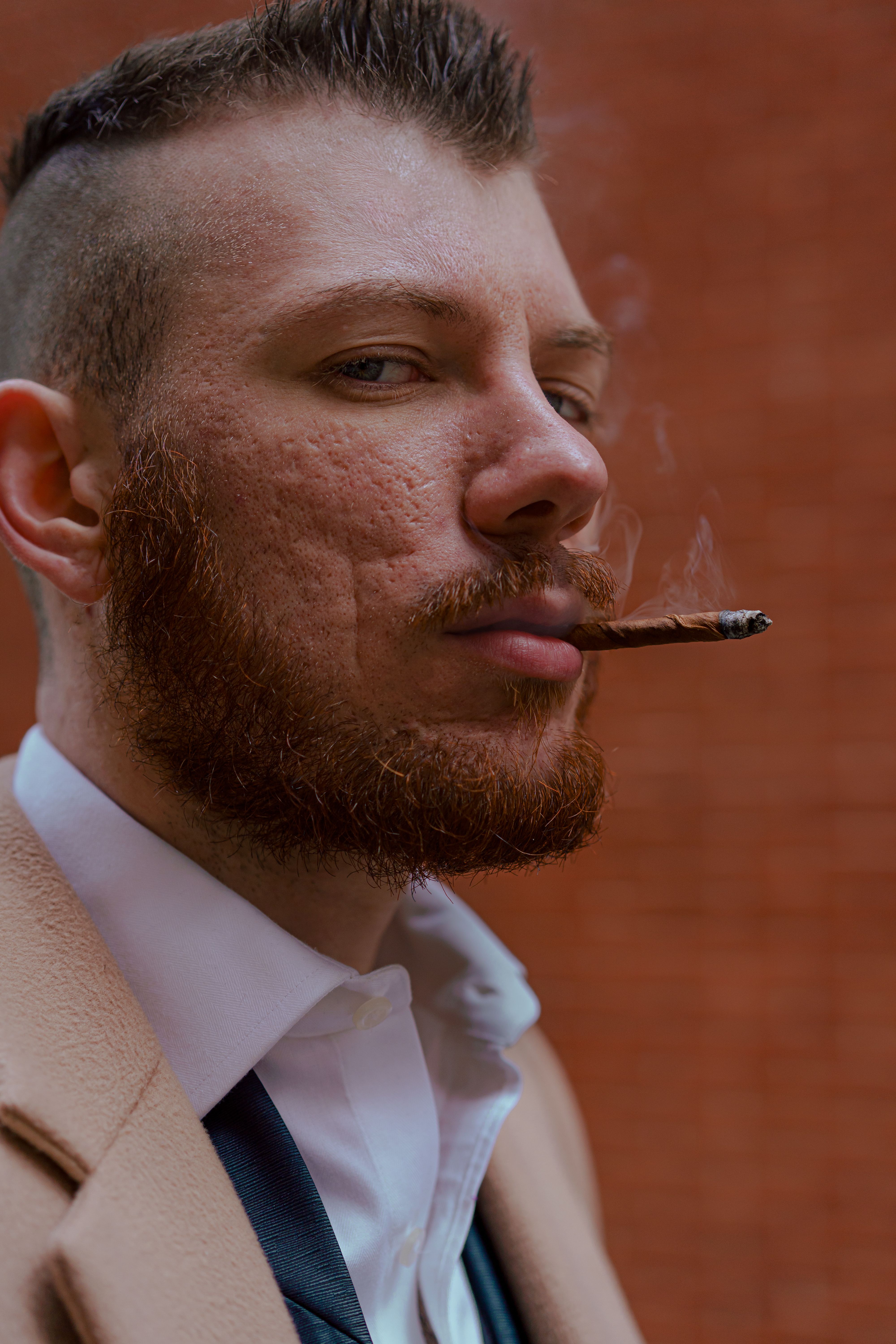 Portrait of a bearded man smoking a cigar in a suit against a brick wall.