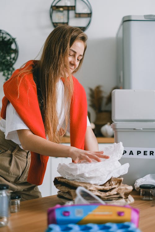 Smiling responsible female with long hair putting paper in heap while sorting trash in kitchen