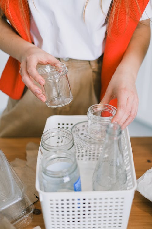 Crop anonymous female in casual clothes putting glass jars and bottles into plastic container while sorting waste in kitchen