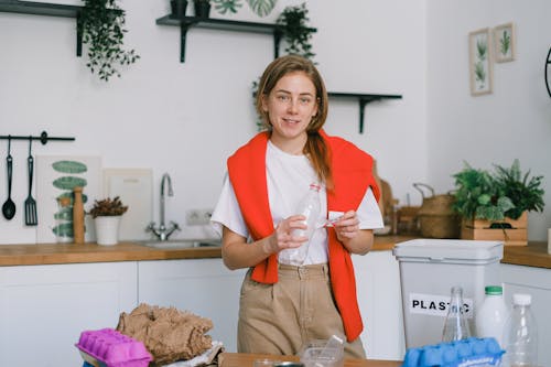 Female holding plastic bottle and looking at camera while sorting trash in blurred kitchen