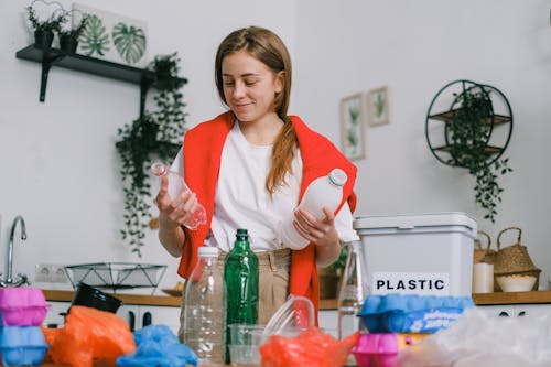 Cheerful female in casual clothes standing with plastic bottles in hands while sorting trash in light kitchen in daytime