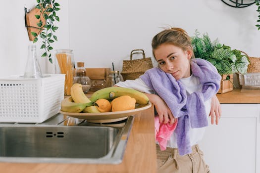 Positive young woman in casual clothes leaning on wooden counter near plate with fresh fruits and tableware in light kitchen in apartment