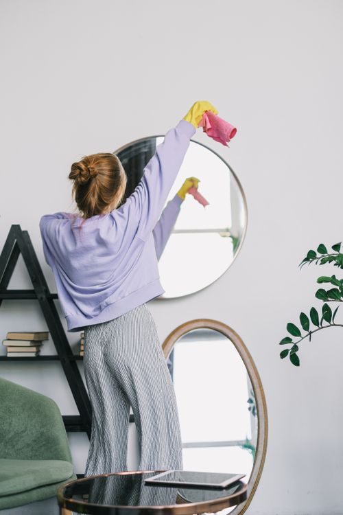 Free A woman cleans a wall mirror wearing casual attire in a stylish indoor setting, emphasizing cleanliness. Stock Photo