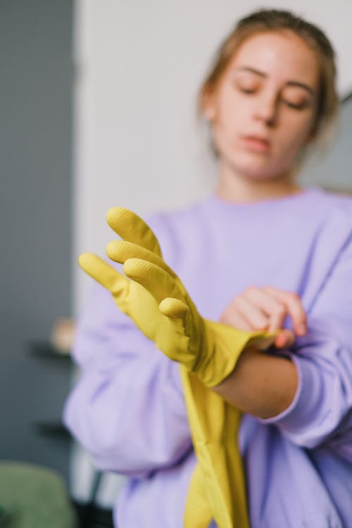 Calm blurred young female in casual clothes reaching out hand and wearing latex gloves in light room