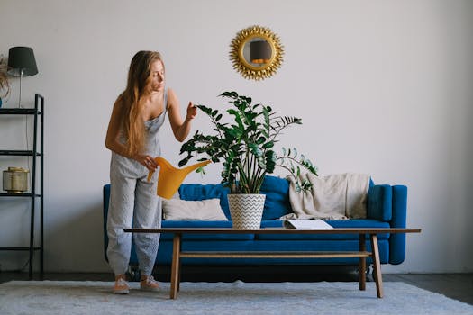 Full body of lady standing at couch while watering plant in pot with can on table at home