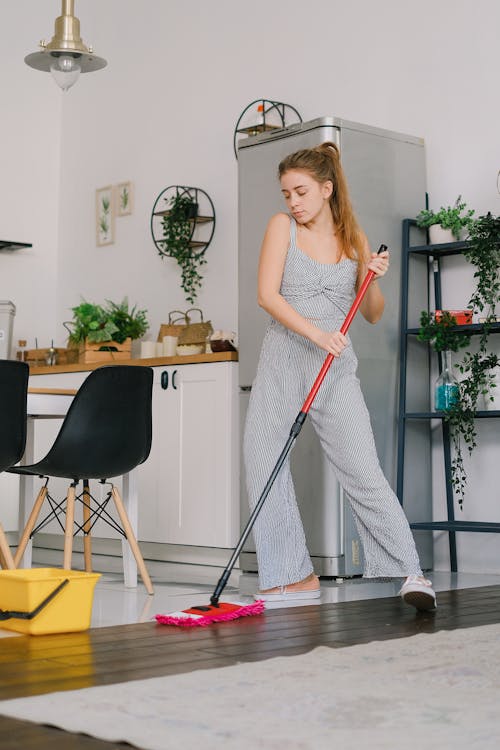 Free Young carefree female in jumpsuit dancing while cleaning parquet with mop in light house room Stock Photo