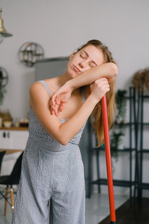 Free Sleepy young woman with closed eyes in jumpsuit leaning on handle of mop in light house room Stock Photo