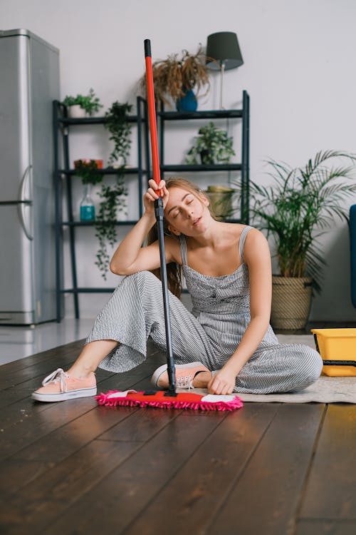Free Exhausted young female with closed eyes leaning on handle of mop while sitting on carpet after washing floor in house Stock Photo