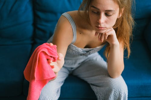 Crop exhausted young female with long hair in casual clothes leaning on hand while sitting on couch with dusting cloth during household routine