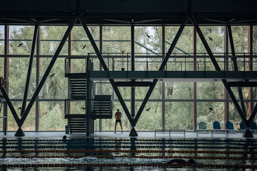 Anonymous shirtless male athlete exercising under balcony while reflecting in swimming pool in building with decorative wall