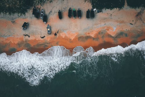 Aerial view of powerful sea with foamy waves washing sandy beach with moored wooden boats in tropical country