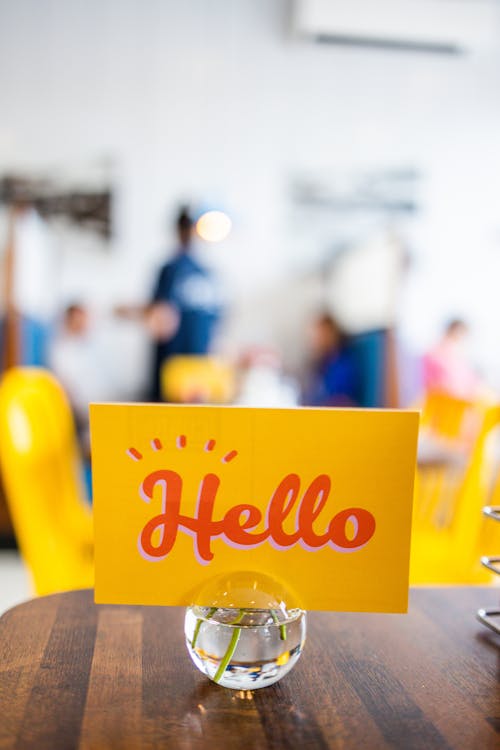 Yellow signboard with Hello title on glass vase with stems in water on table in cafeteria