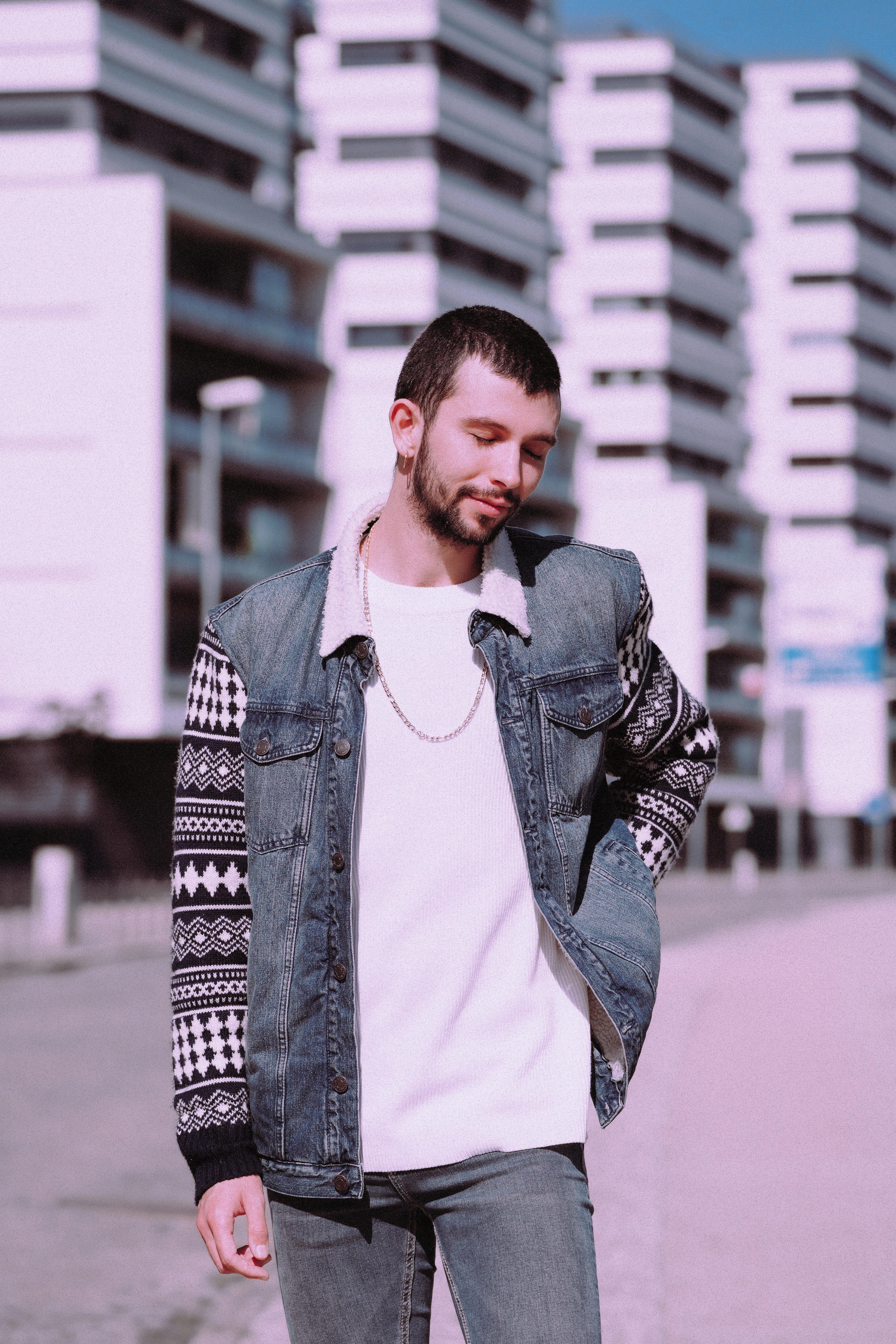 Stylish young man wearing denim and patterned sweater, standing confidently outdoors in urban area.