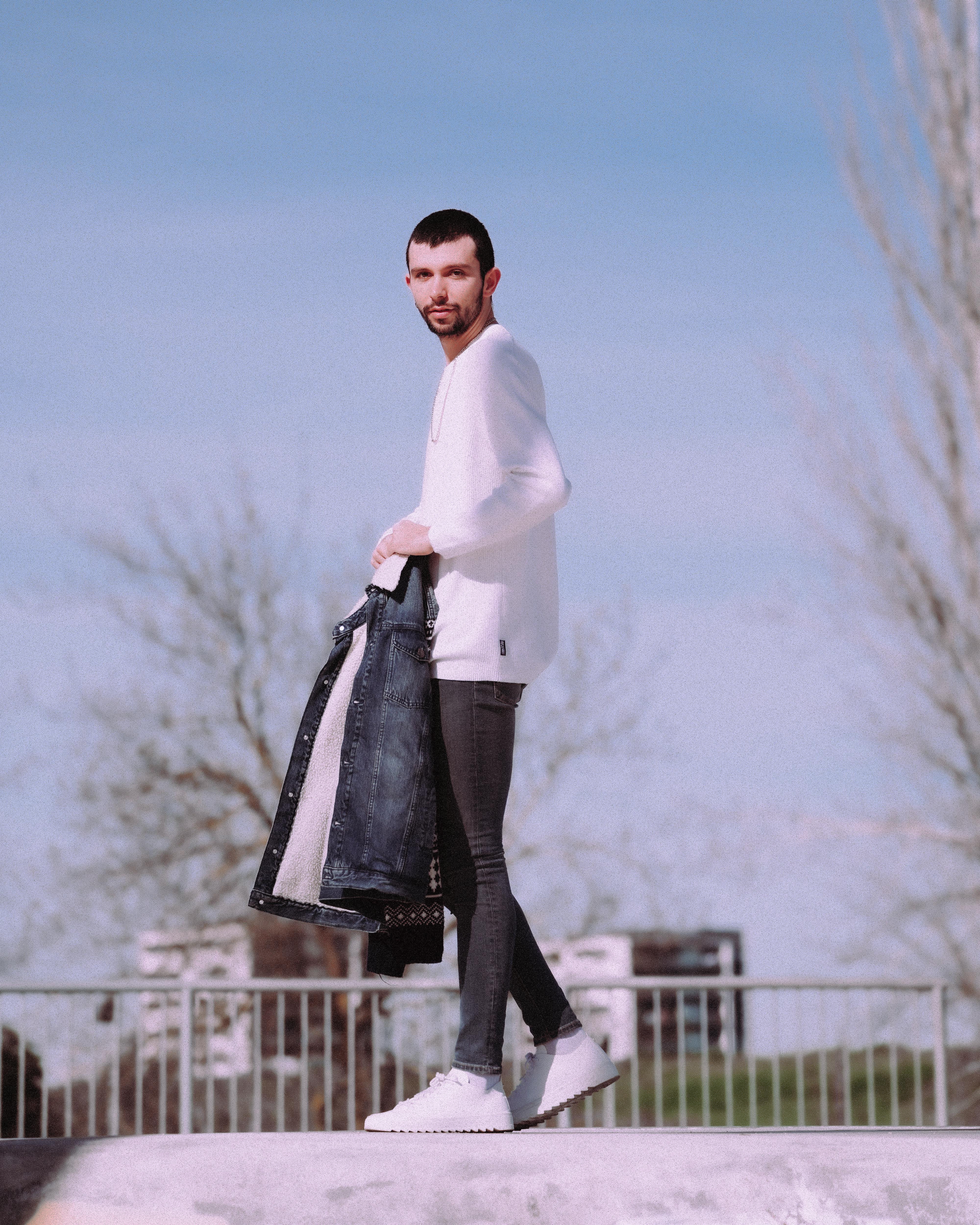Man holding denim jacket in an urban area, showcasing modern fashion style on a sunny day.