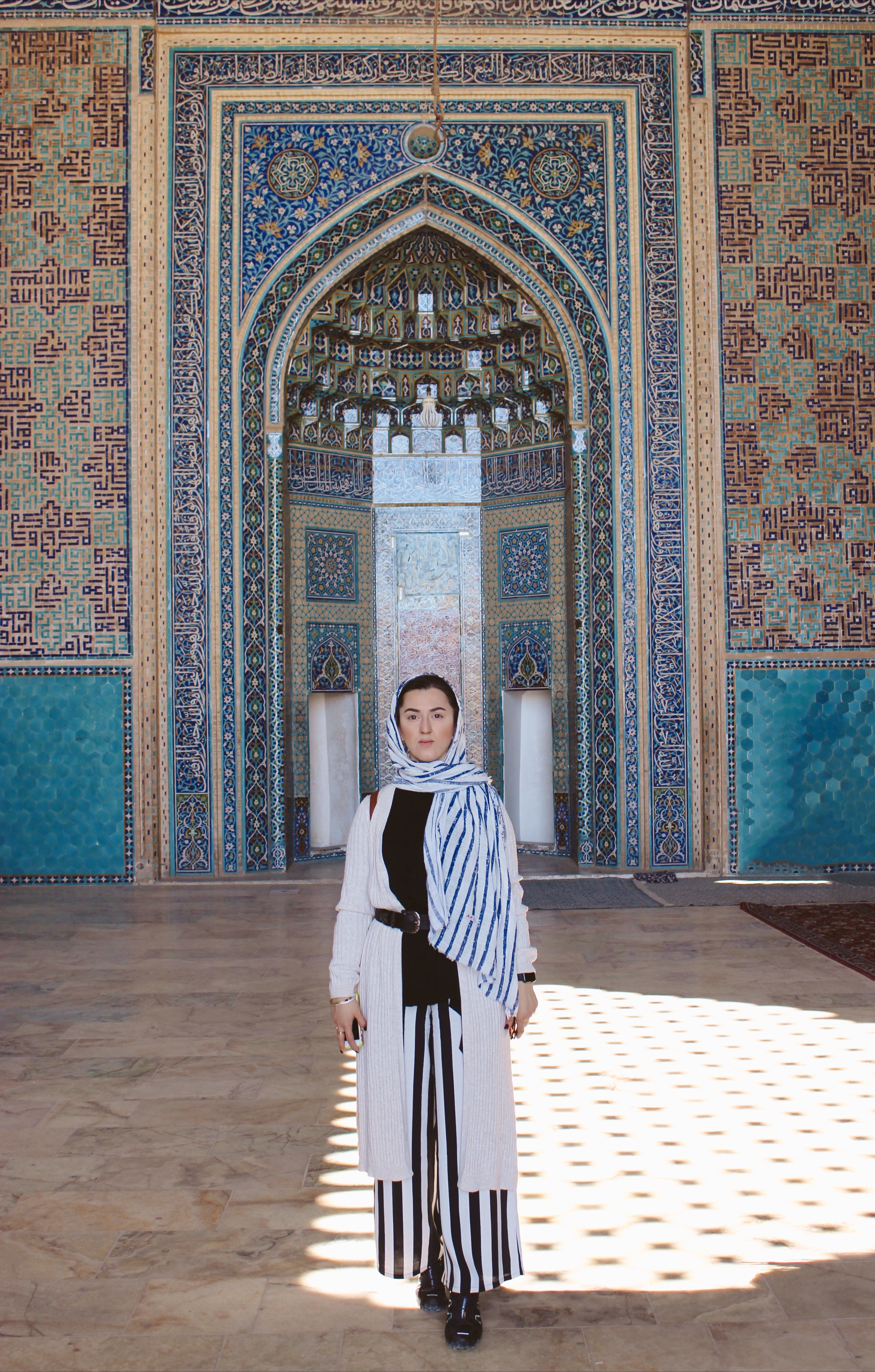 A woman in a hijab stands gracefully within a beautiful, intricately decorated architectural setting.