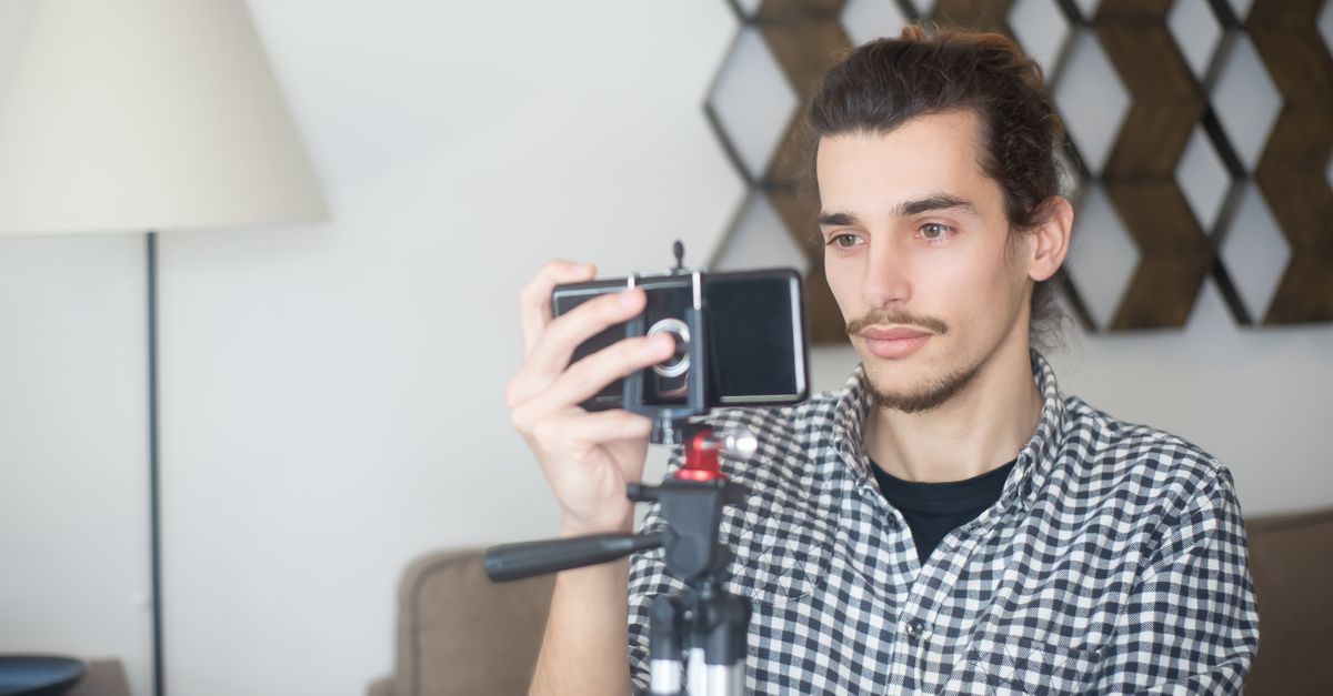 Young man with plaid shirt using a smartphone and tripod to create content indoors in Portugal.