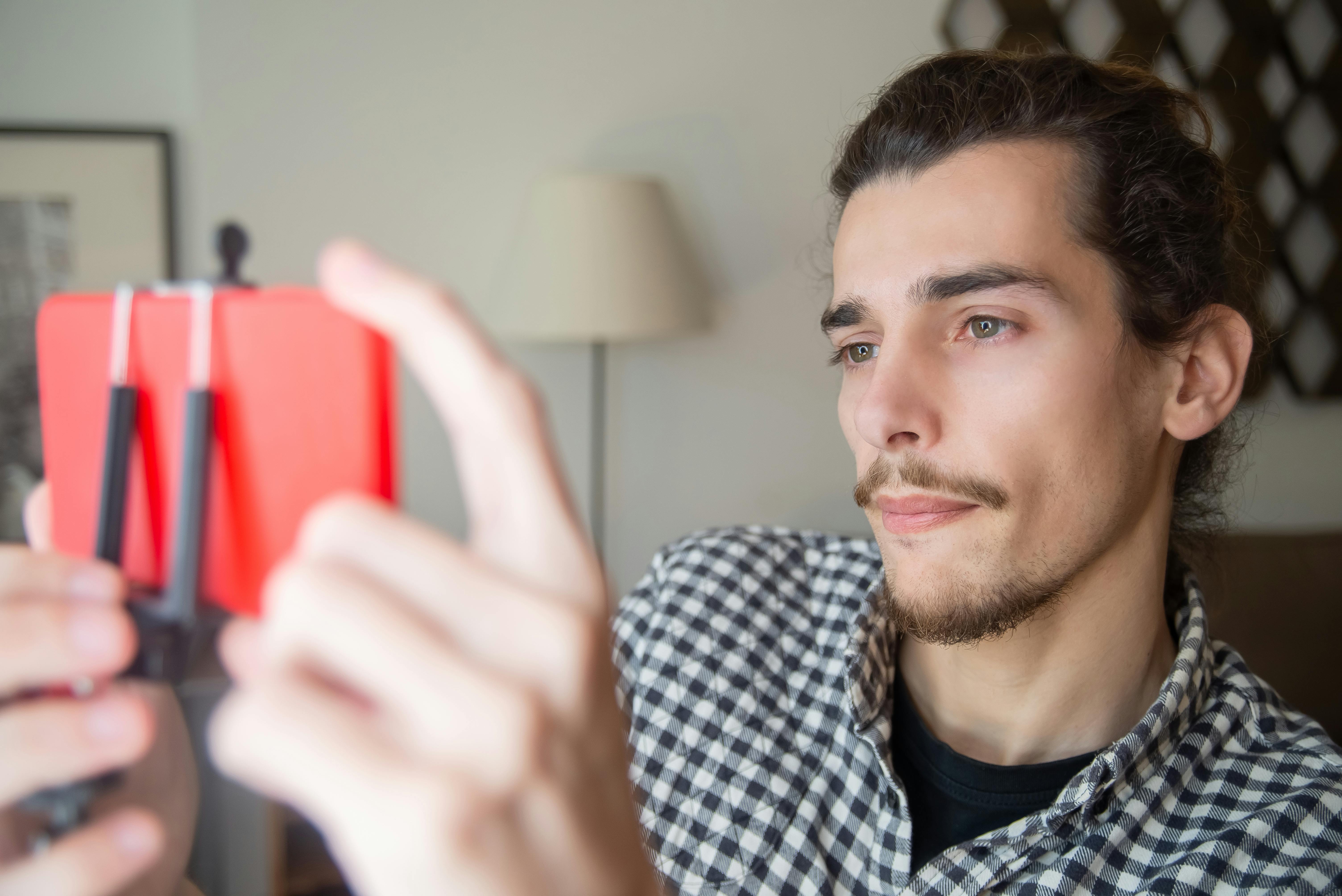 A young bearded man in a checkered shirt live streaming indoors using a smartphone on a tripod