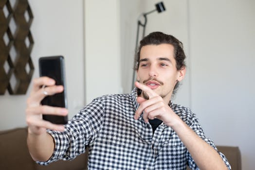Young man in checkered shirt live streaming or vlogging indoors using smartphone.