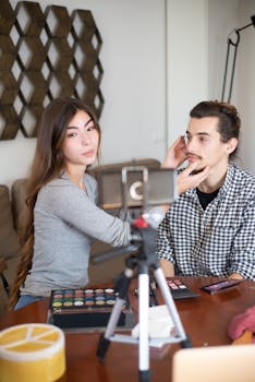 Asian woman applies makeup to a Caucasian man for a vlogging session, filmed using a tripod.