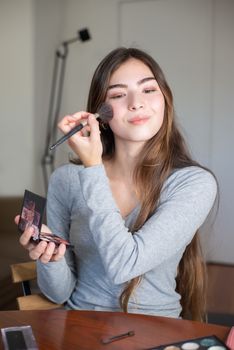A young woman cheerfully applying makeup with a brush indoors.