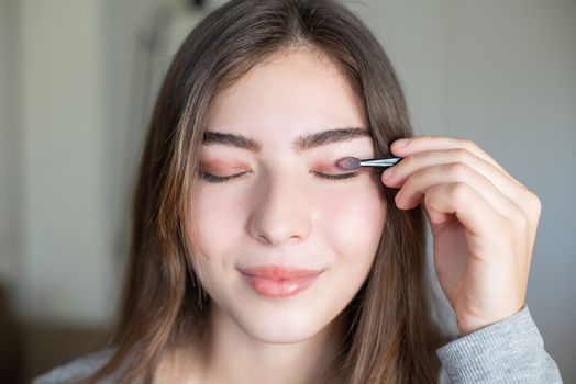 Close-up of a young woman applying eyeshadow with eyes closed indoors.