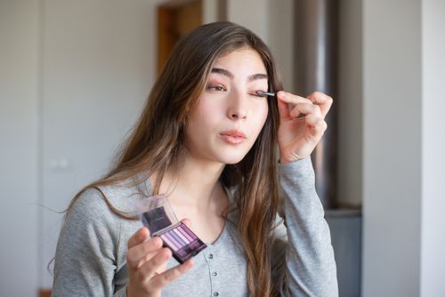 Free Asian woman applying makeup with eyeshadow palette indoors, showcasing beauty routine. Stock Photo
