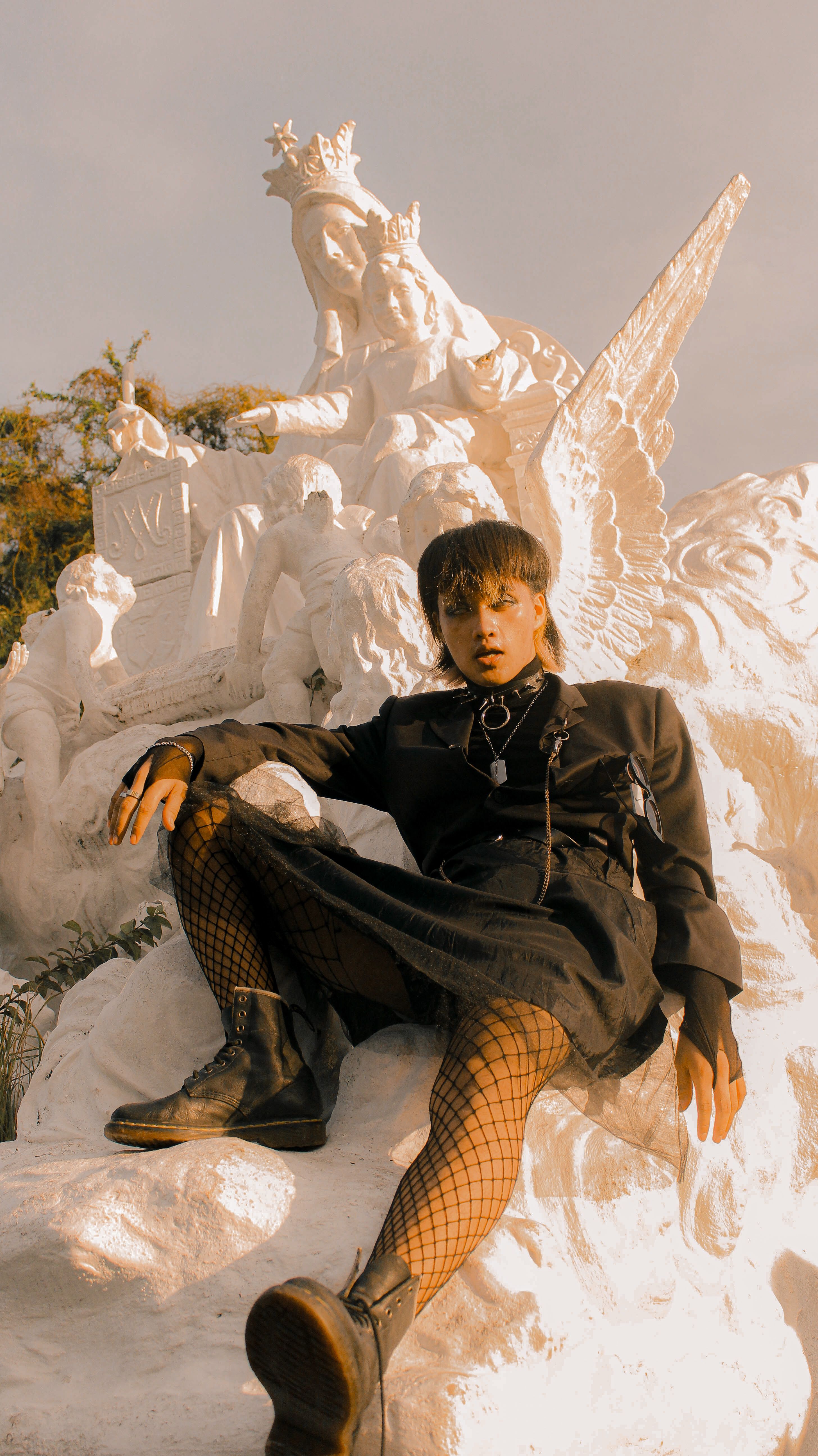 Stylish person dressed in gothic attire sitting on a religious statue outdoors during the day.