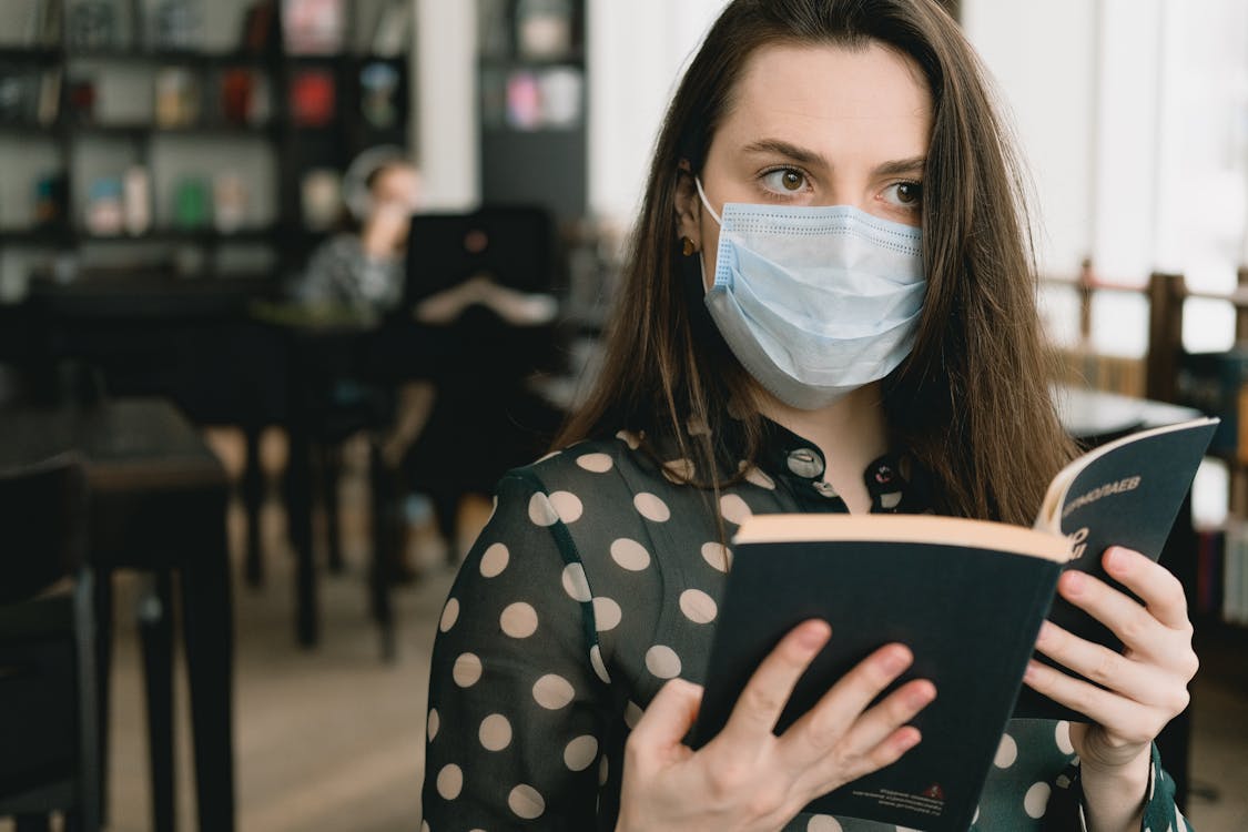 Free Young woman in protective mask reading book in library Stock Photo
