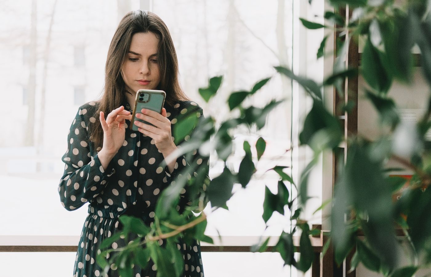Woman in polka dot dress using smartphone indoors, surrounded by plants.