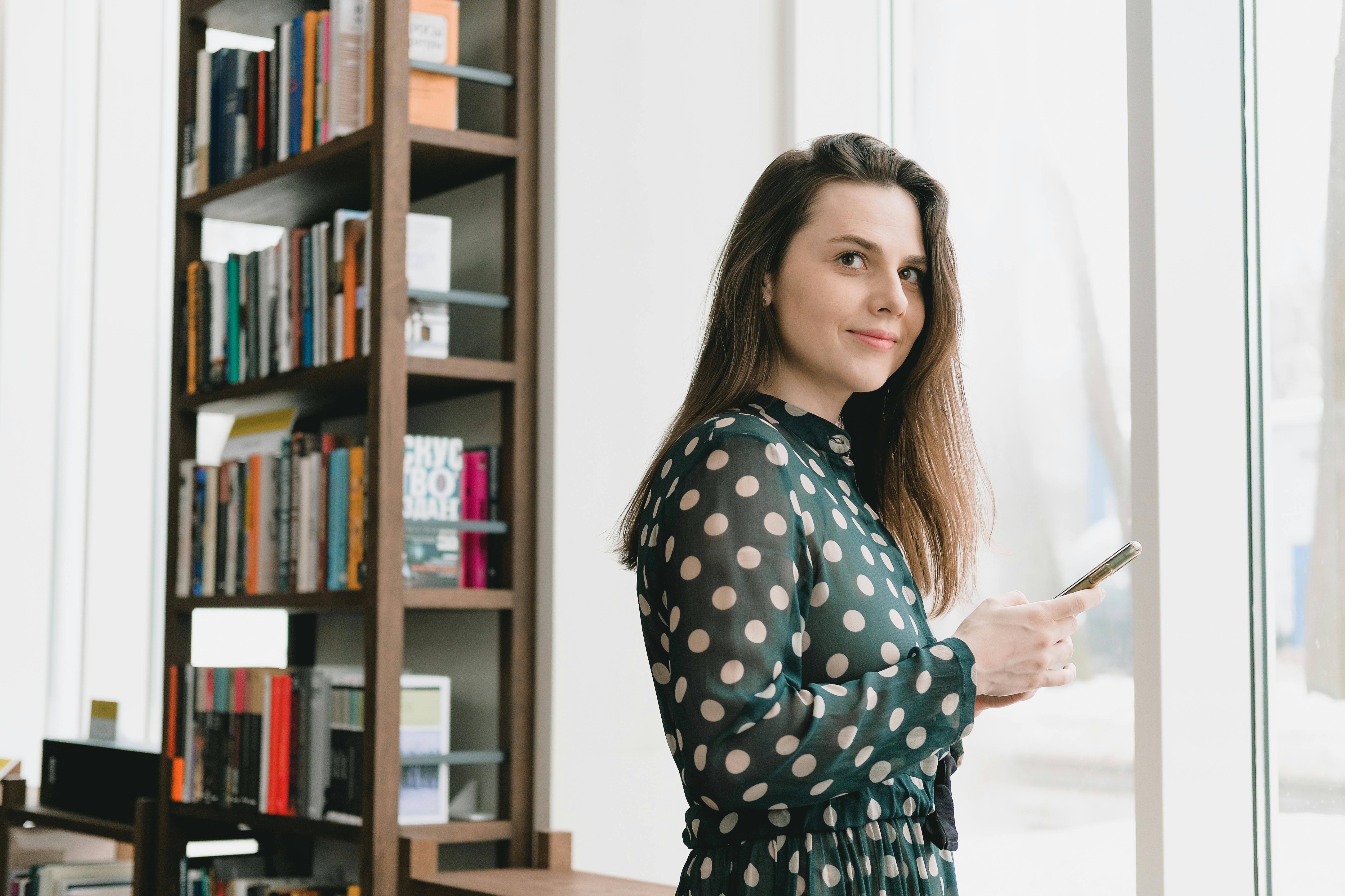 smiling young lady messaging on smartphone near window in library