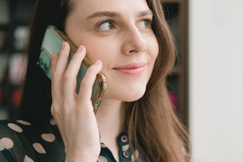 Free Crop young happy female millennial with long dark hair smiling and looking away while having phone conversation near window on sunny day Stock Photo