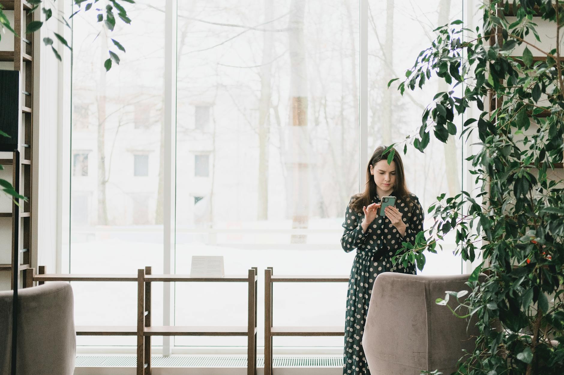 Concentrated young lady with long hair in stylish dress standing near panoramic windows and messaging on mobile phone on snowy winter day in modern workspace