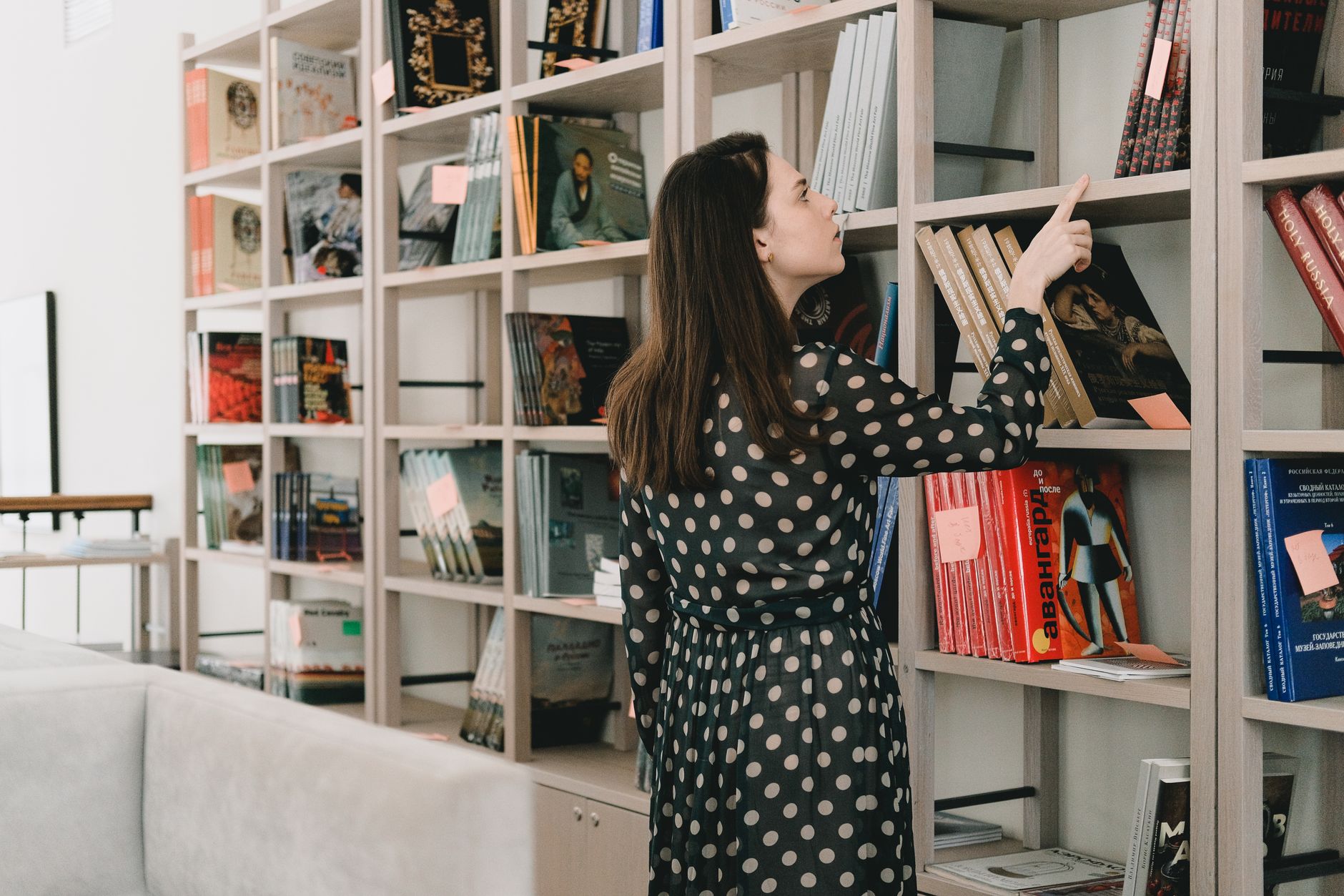 Back view of young female standing near bookcase and selecting literature for leisure