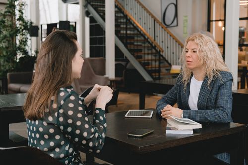 Free Businesswomen sitting at table with book and devices and having conversation about project in modern workplace Stock Photo