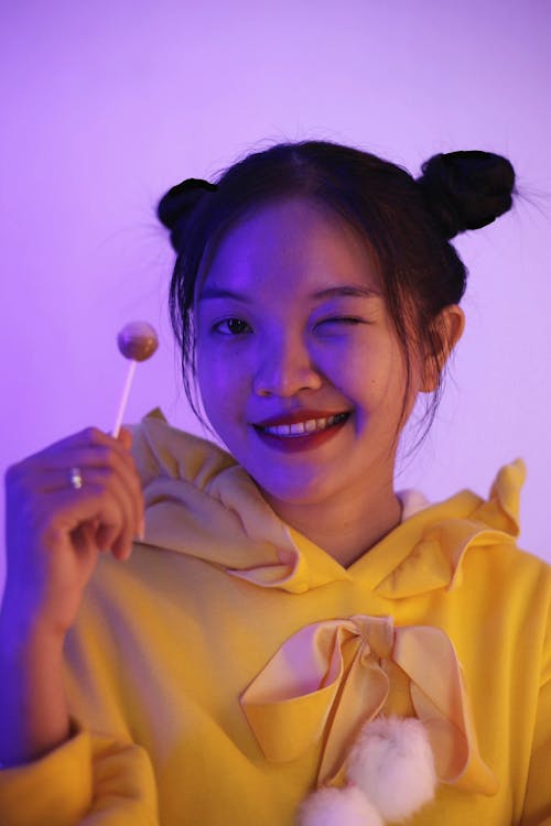 Positive young Asian female with lollipop in hand winking and looking at camera while standing on purple background in studio