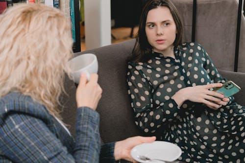 Free Young female friends in elegant outfits sitting on comfortable sofa and talking during coffee break in cozy cafe Stock Photo