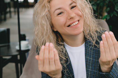 Free Crop cheerful adult female with curly blond hair smiling and gesticulating at camera while sitting in armchair in cafe and recording vlog Stock Photo