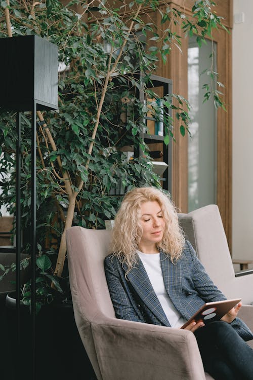 Confident adult female manager with curly blond hair in smart casual outfit sitting in comfortable armchair and reading notes on tablet in modern office