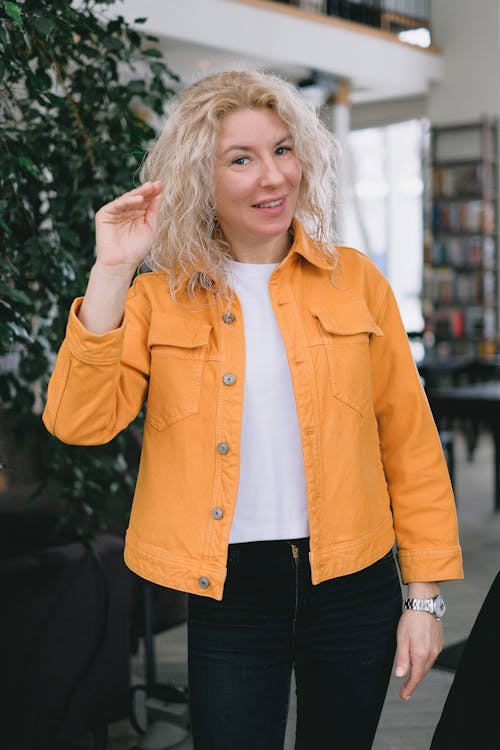 Positive adult female blogger with blond hair in casual clothes standing in light room and smiling while recording vlog