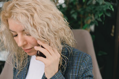 Crop concentrated female entrepreneur with curly blond hair in formal outfit sitting in armchair and looking down attentively while having phone conversation during work in modern office