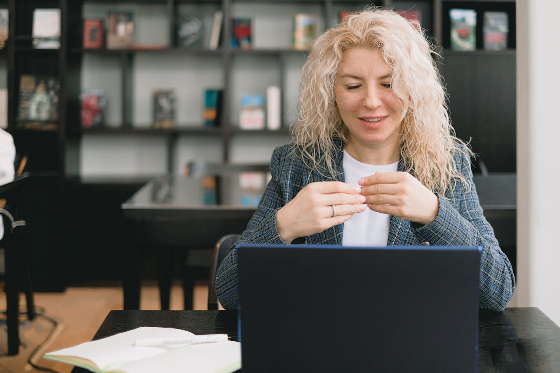 Positive young female employee with curly blond hair in elegant clothes smiling while having video call via laptop sitting at table with notebook in modern office